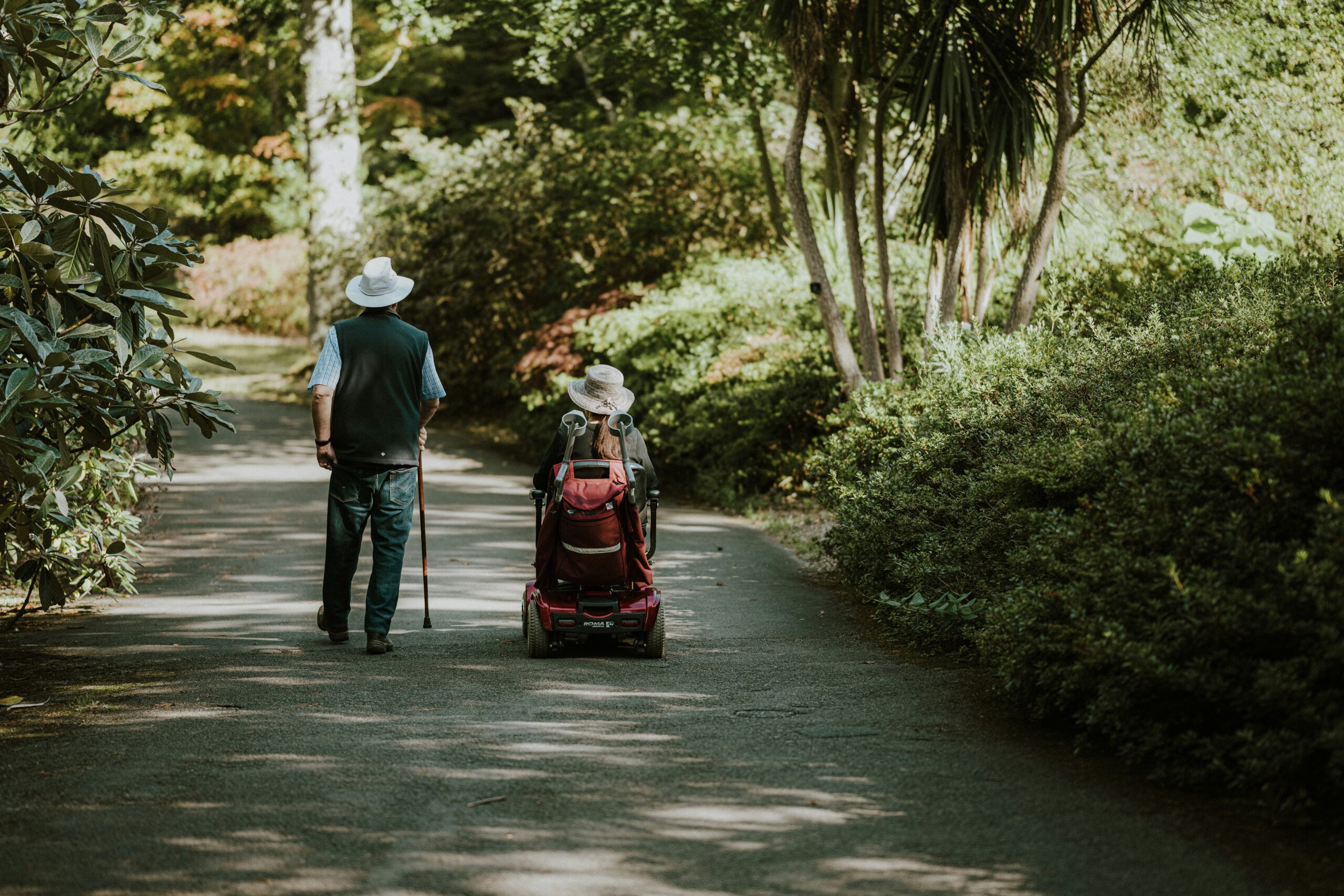 annie-spratt-A2bvXVD5qgo-unsplash A photo of an older lady in a mobility scooter with her husband walking with a cane through a park.